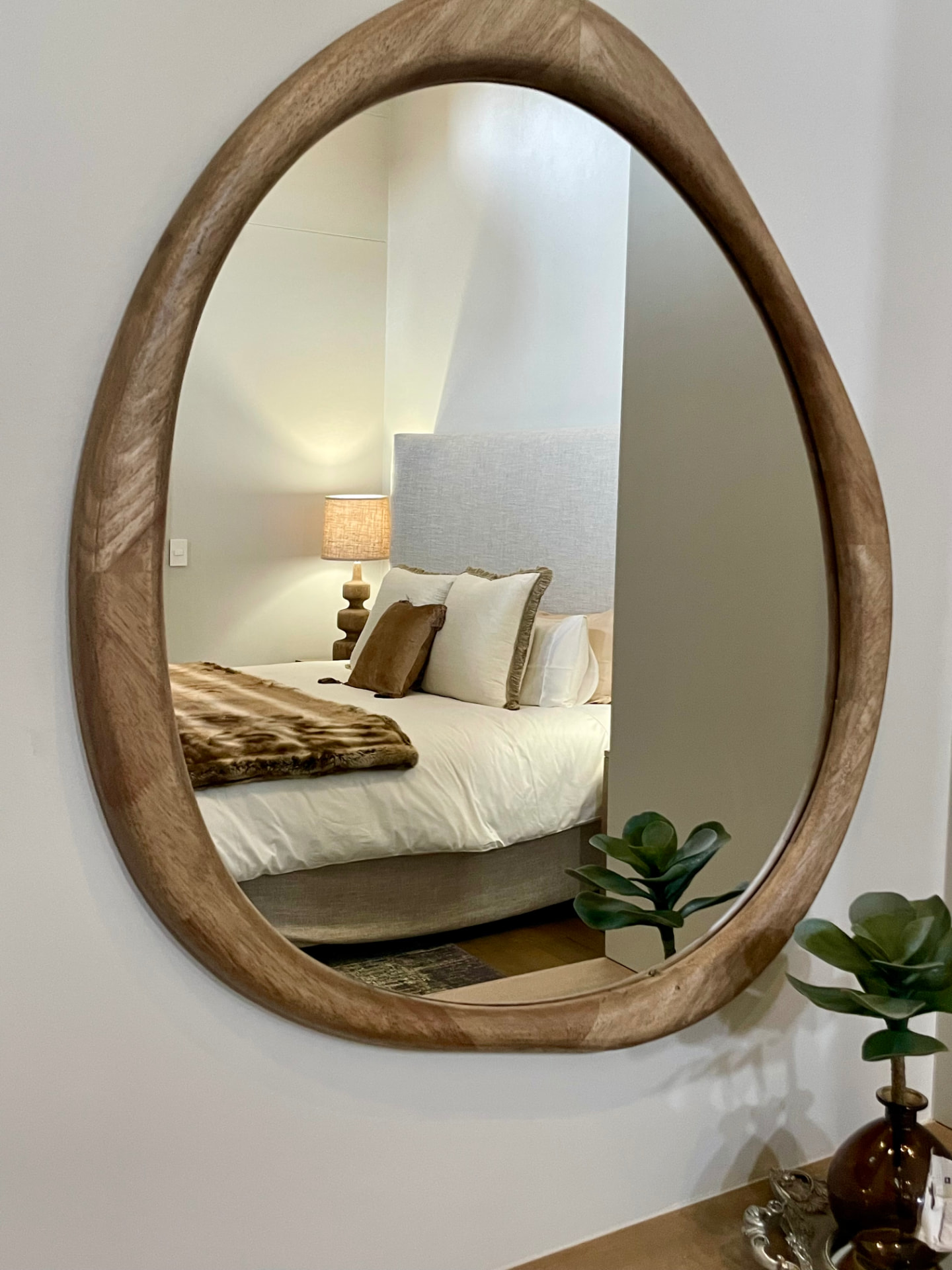 Organic timber-framed mirror reflecting a bedroom with layered linen and fur throw